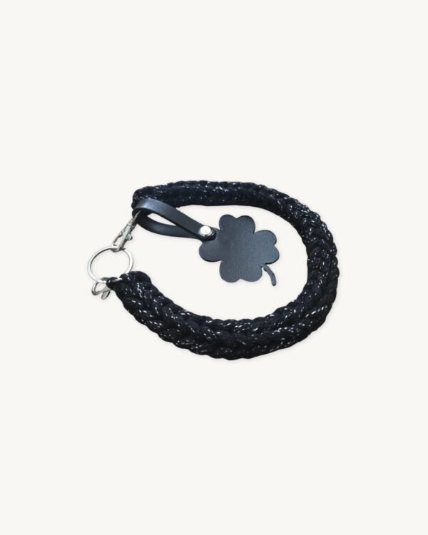 Angled side profile of the CHIU STYLE clover vegan leather braided keychain in black with silver thread resting on a natural wood surface, highlighting texture and clover tag