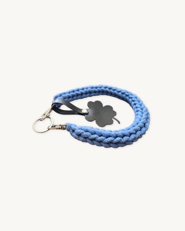 Angled side profile of the CHIU STYLE clover vegan leather braided keychain in cornflower blue resting on a natural wood surface, highlighting texture and clover tag